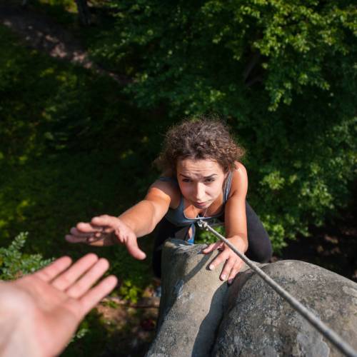 Young fearless woman climbing on large boulder, her male partner giving her a hand. Helping hand. View from the top